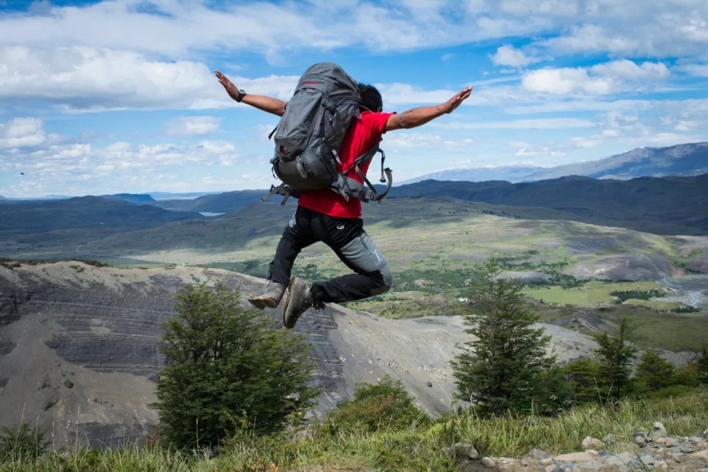 Hiker with a gray backpack and red shirt leaps with arms outstretched above a Patagonian valley and distant mountains