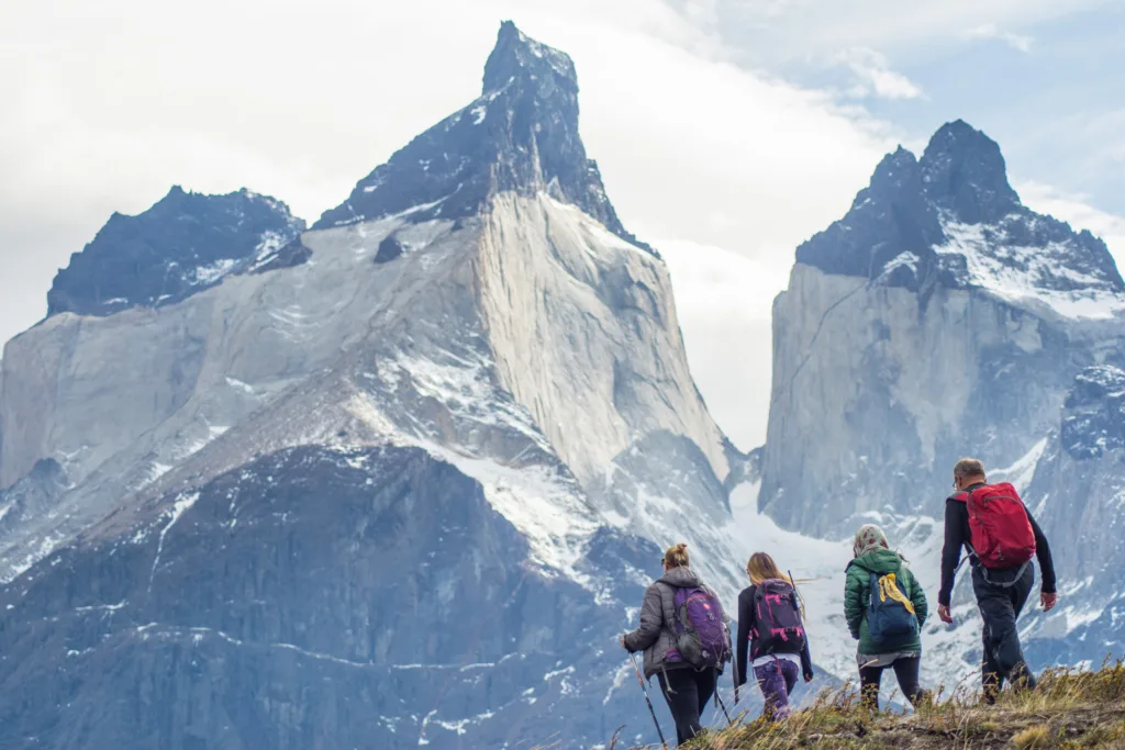 Four hikers with backpacks and trekking poles walk along a ridge toward towering snow-dusted granite peaks