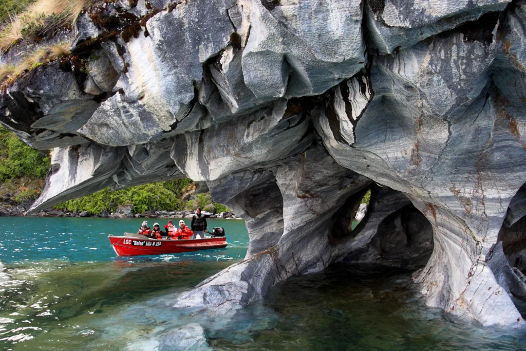 Red boat with passengers in life jackets beside swirled gray and blue marble caves on a turquoise lake